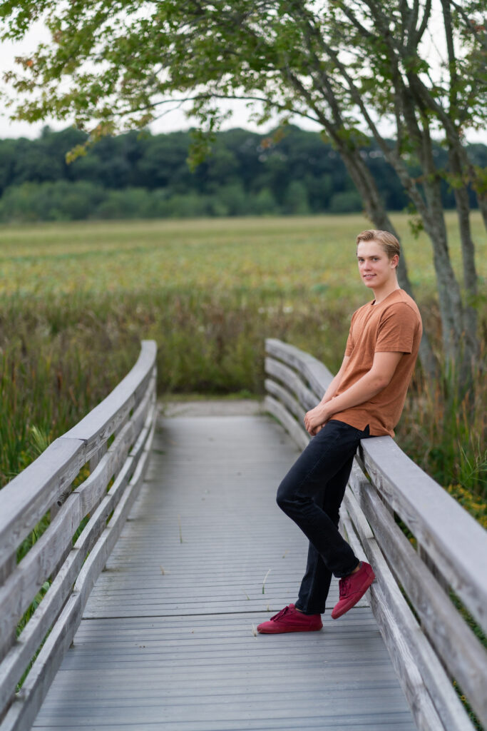 Great Meadows in Concord, MA is a senior portrait location with lots of nature. This portrait is of a high school senior boy standing on a wooden bridge footpath. 