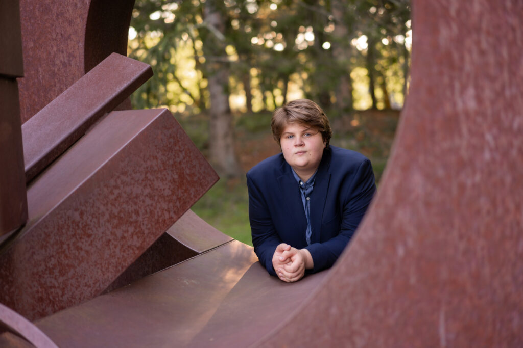 Senior portrait of a boy outdoors with an art sculpture. Photographed at the deCordova museum in Lincoln, MA by Pierre Chiha Photographers. The boy is dressed nicely in a button down shirt and jacket. 
