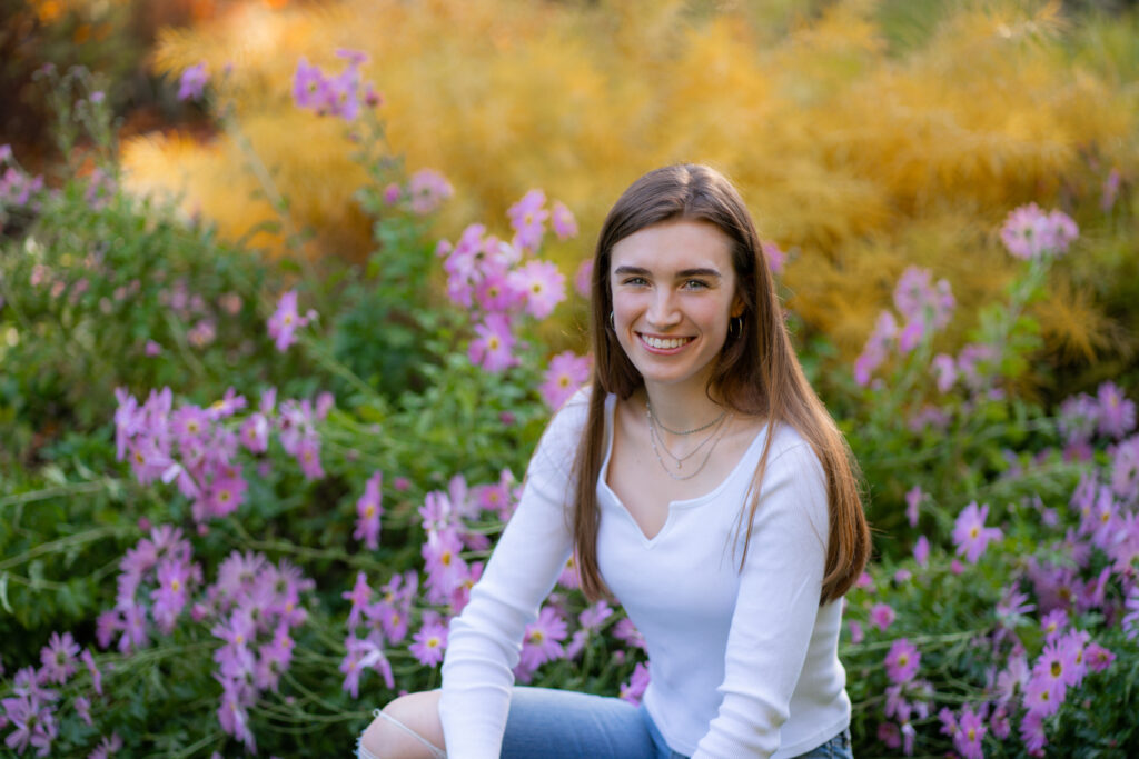 Senior portrait location at this girl's home backyard. She is kneeling next to bushes of flowers wearing a long sleeve white shirt. Photographed by Pierre Chiha Photographers